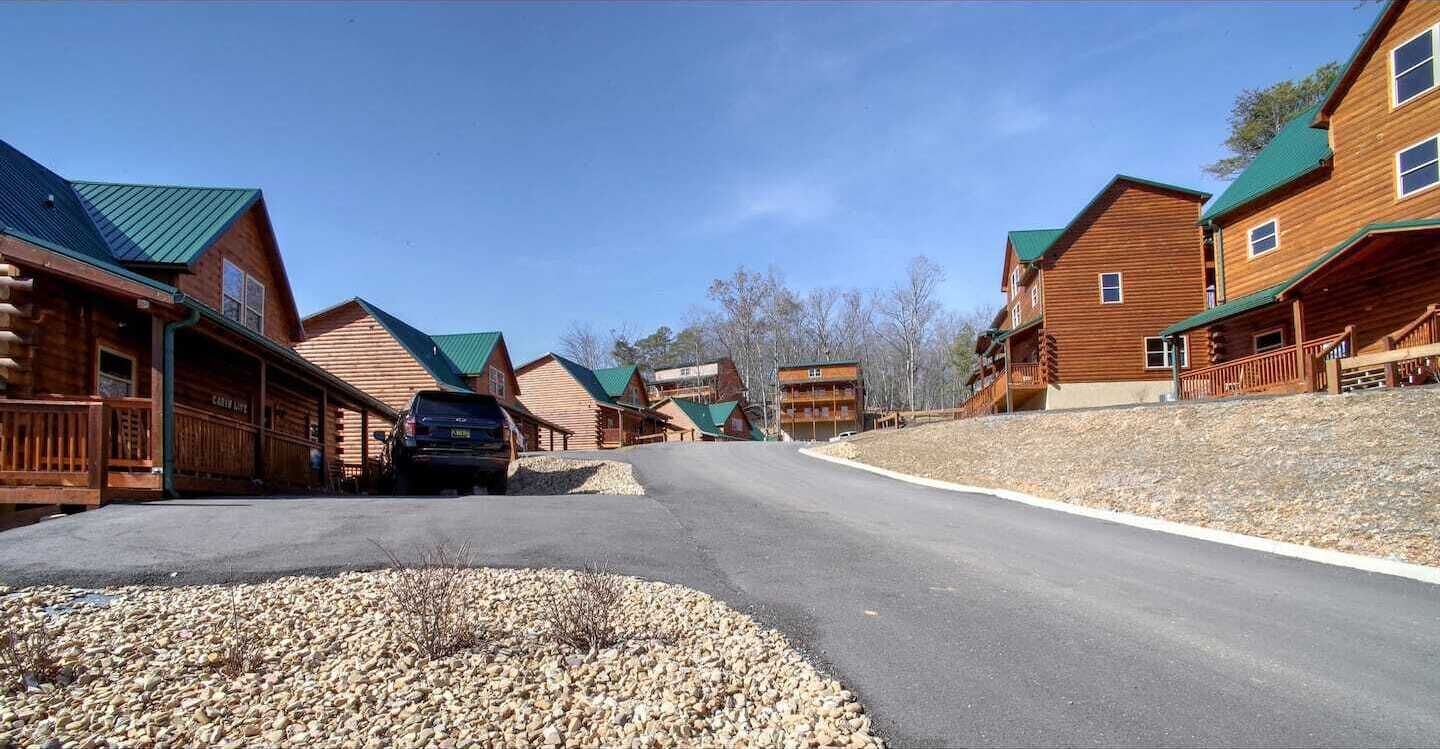 A row of log cabins with a paved road in front, a parked car, and a gravel area.