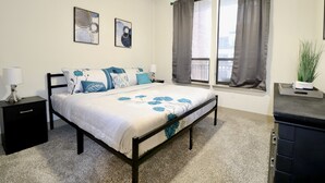 Quarto com cama, mesa de cabeceira, janela com cortinas e uma planta.