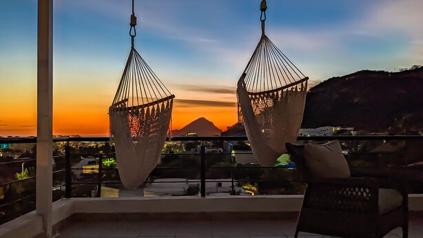 A balcony with two hanging chairs and a view of a sunset over a mountain.