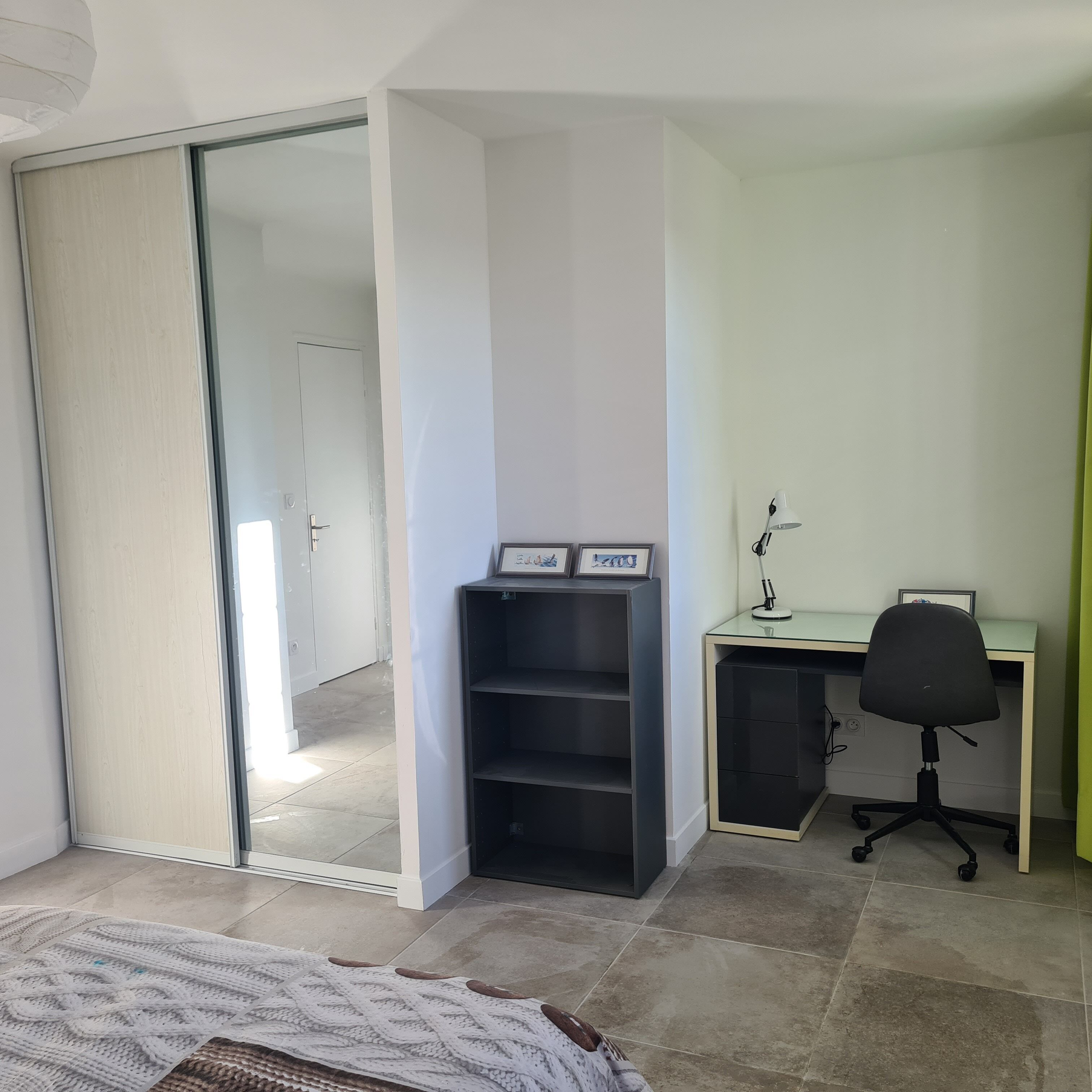 Una habitación con puerta de vidrio corrediza que da a un pasillo, un escritorio con silla y una estantería con cuadros enmarcados.