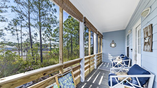 A covered porch with wooden railings, outdoor seating with blue cushions, and a view of pine trees.