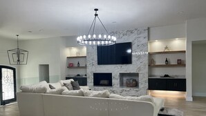 Un salon moderne avec un grand canapé blanc, un téléviseur fixé au mur, un lustre et des étagères intégrées ornées d’objets décoratifs.
