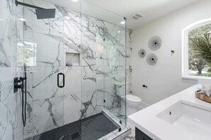A modern bathroom with a glass-enclosed shower, marble walls, a herringbone floor, a white sink, and decorative wall art. - Entire Luxury Home to Enjoy Dallas: Howdy Haven (Dallas)
