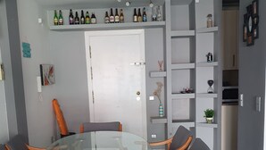A dining area with a glass-top table surrounded by six wooden chairs with grey cushions. Above the table, there is a shelf with various bottles and a decorative light fixture. - Beach apartment (Salou)