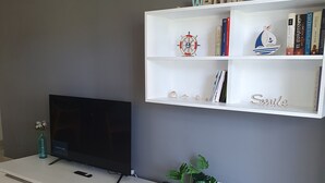 A modern living room with a flat-screen TV on a white platform, a bookshelf with books and decorative items, and a potted plant. - Beach apartment (Salou)