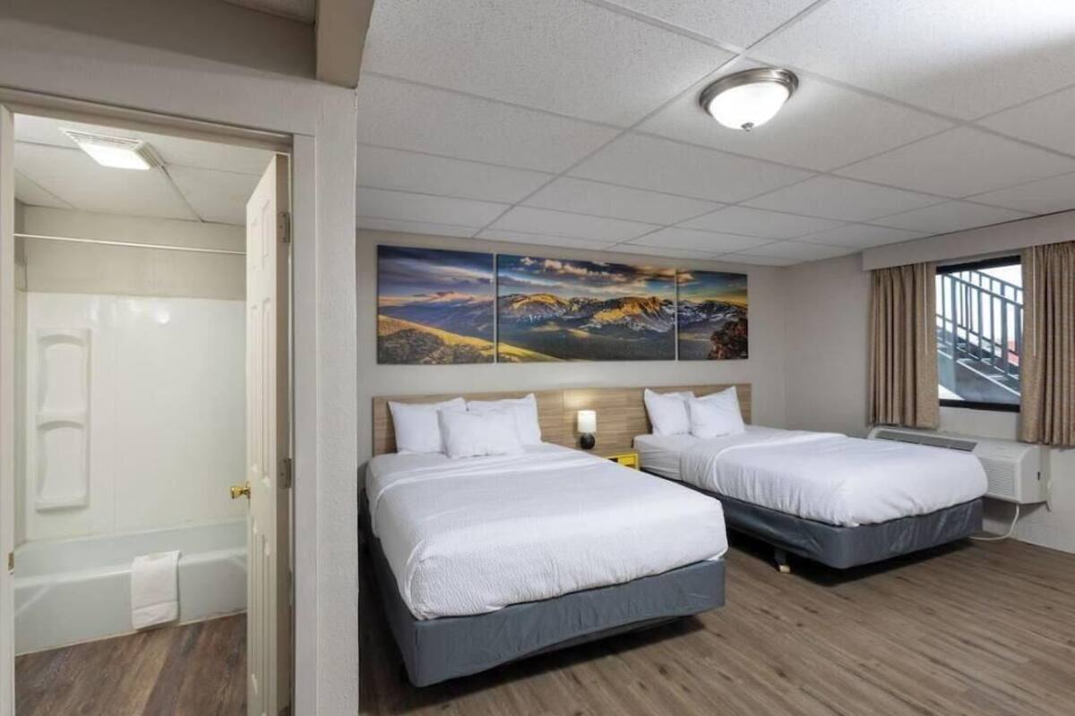A hotel room with two beds, a large window, a painting on the wall, and a bathroom visible through an open door.
