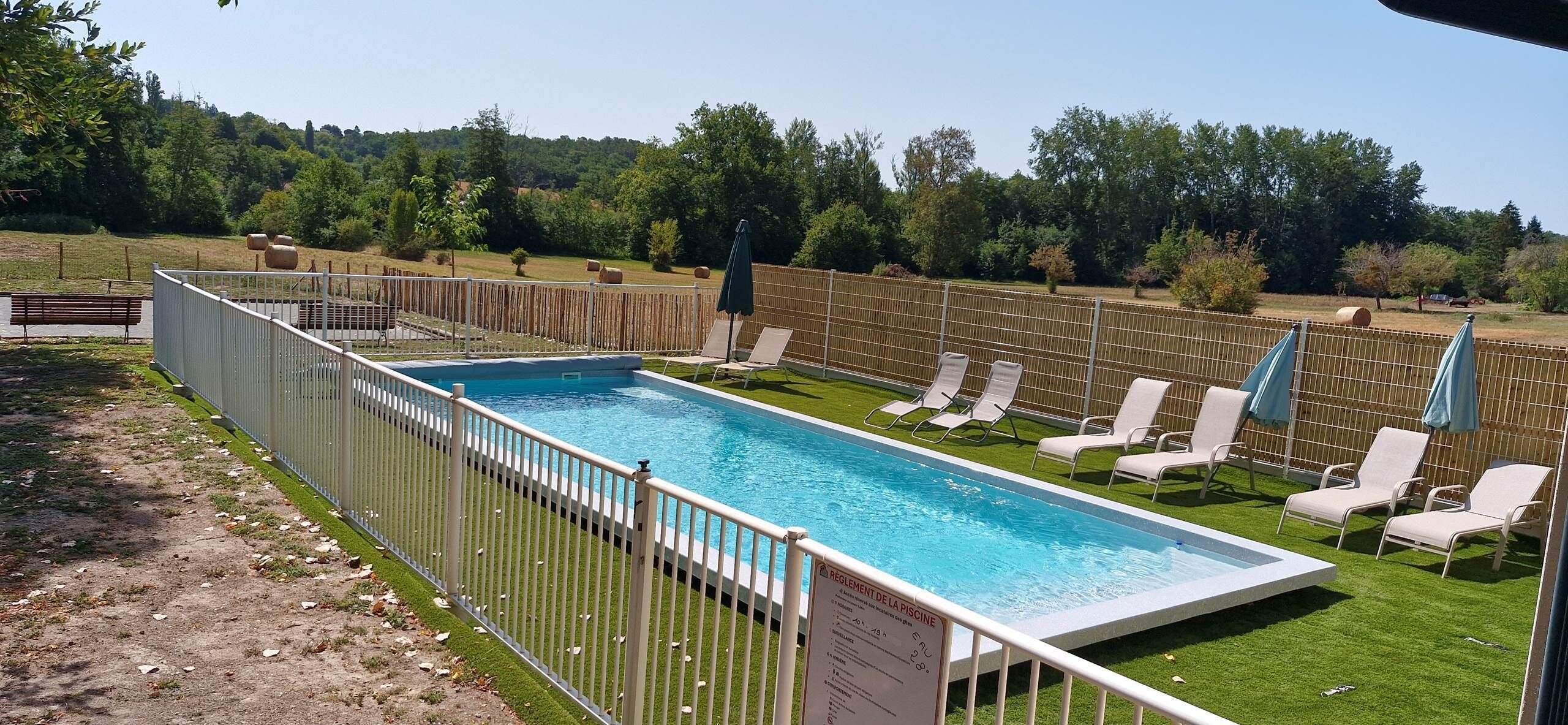 A rectangular swimming pool with a white fence, surrounded by grass and trees.