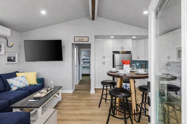 Cocina moderna con armarios blancos, un refrigerador de acero inoxidable y una zona de comedor con mesa de madera y taburetes de bar.