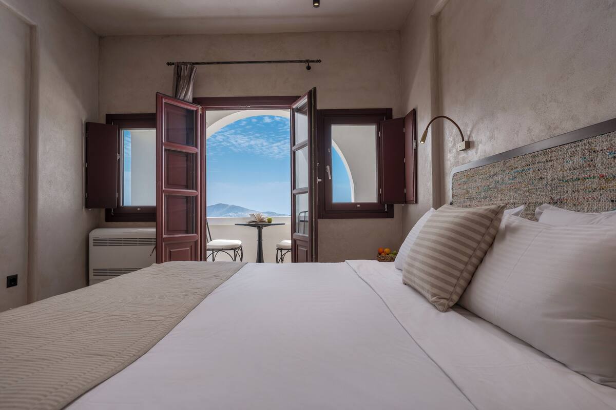 Une chambre avec un grand lit, une vue sur les montagnes par une fenêtre ouverte et une petite table avec des chaises.