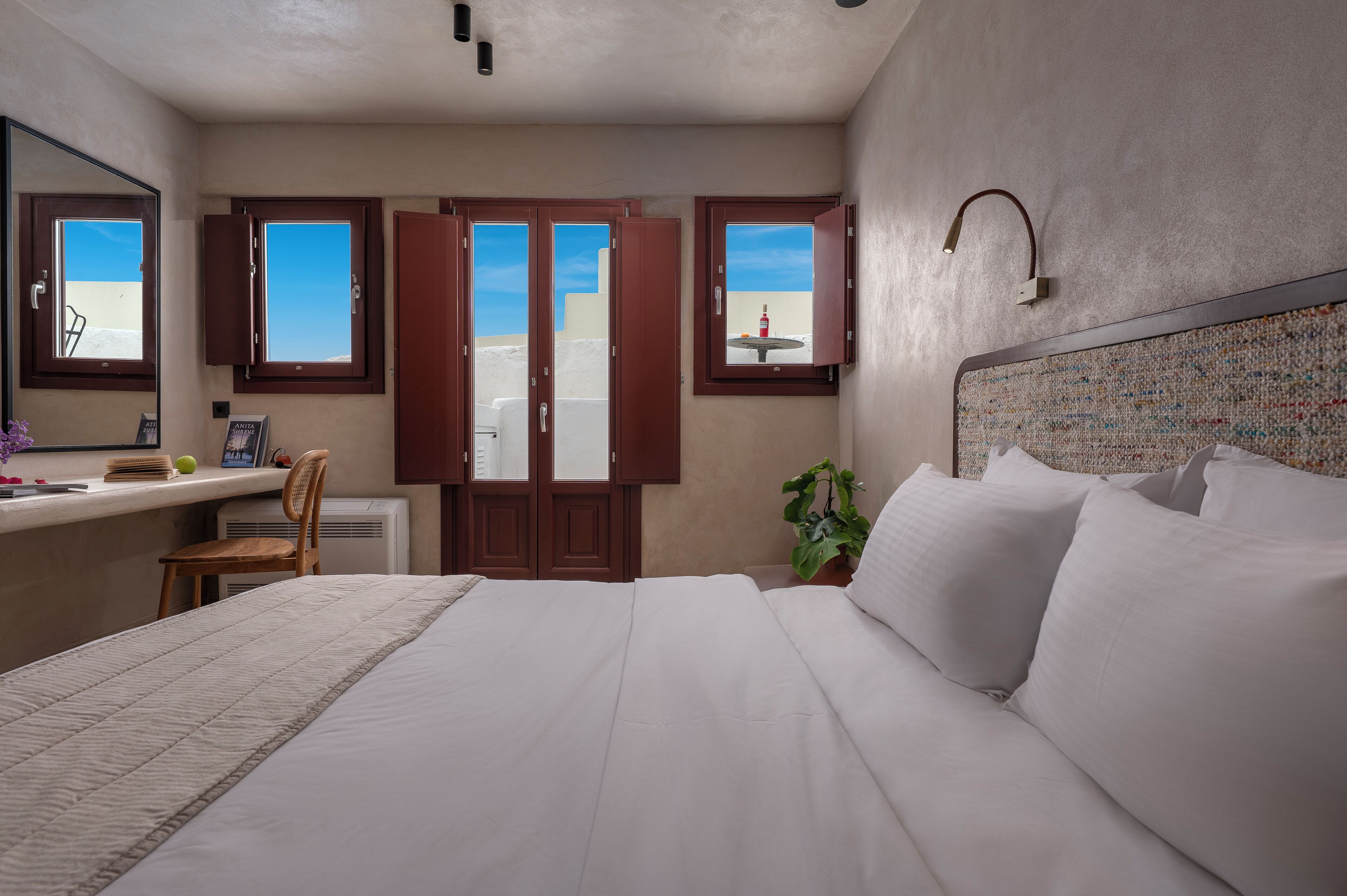 A hotel room with a large bed, a desk with a chair, a small table, and a potted plant.