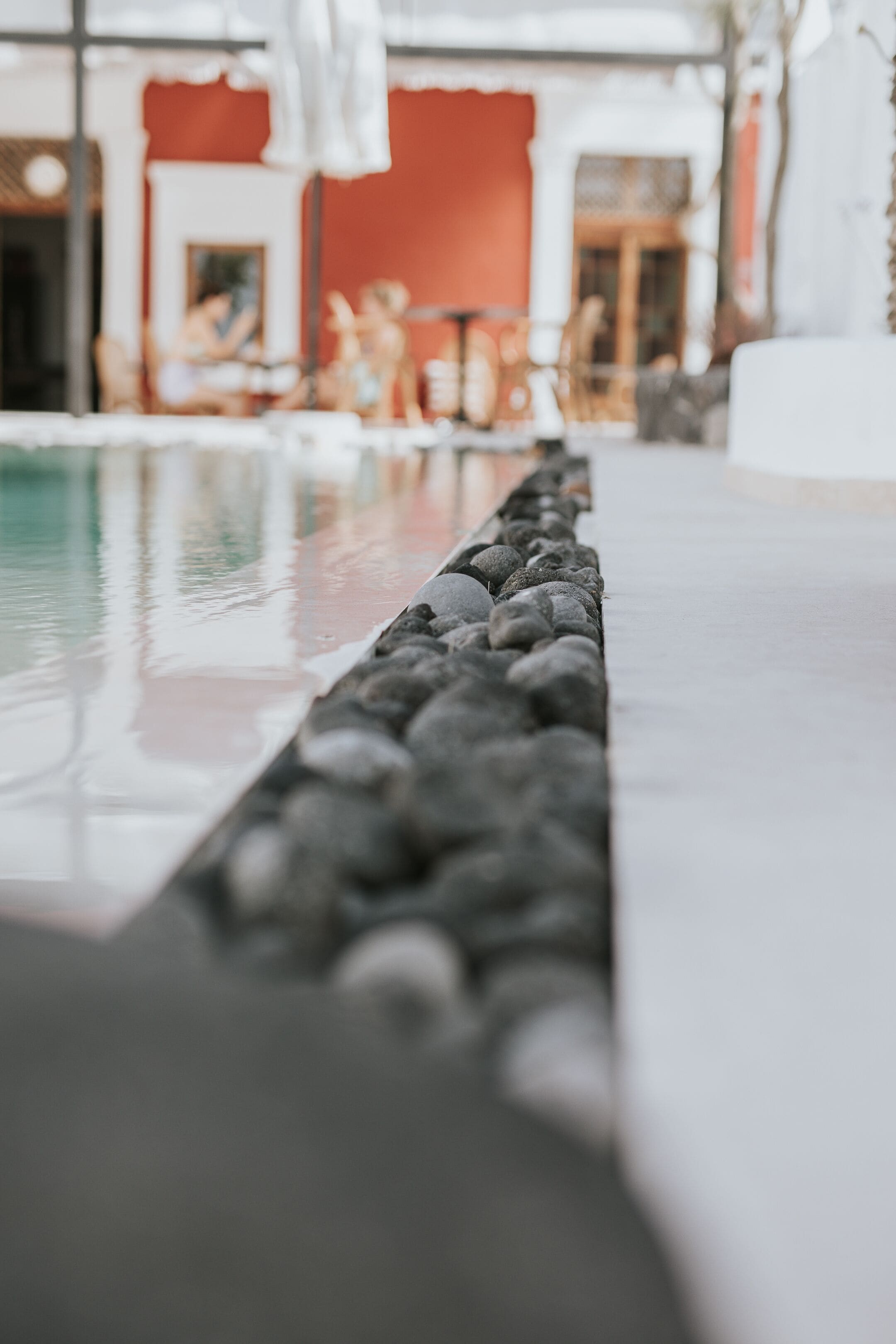 A pool with a pebble-lined edge, a red wall, and outdoor seating in the background.