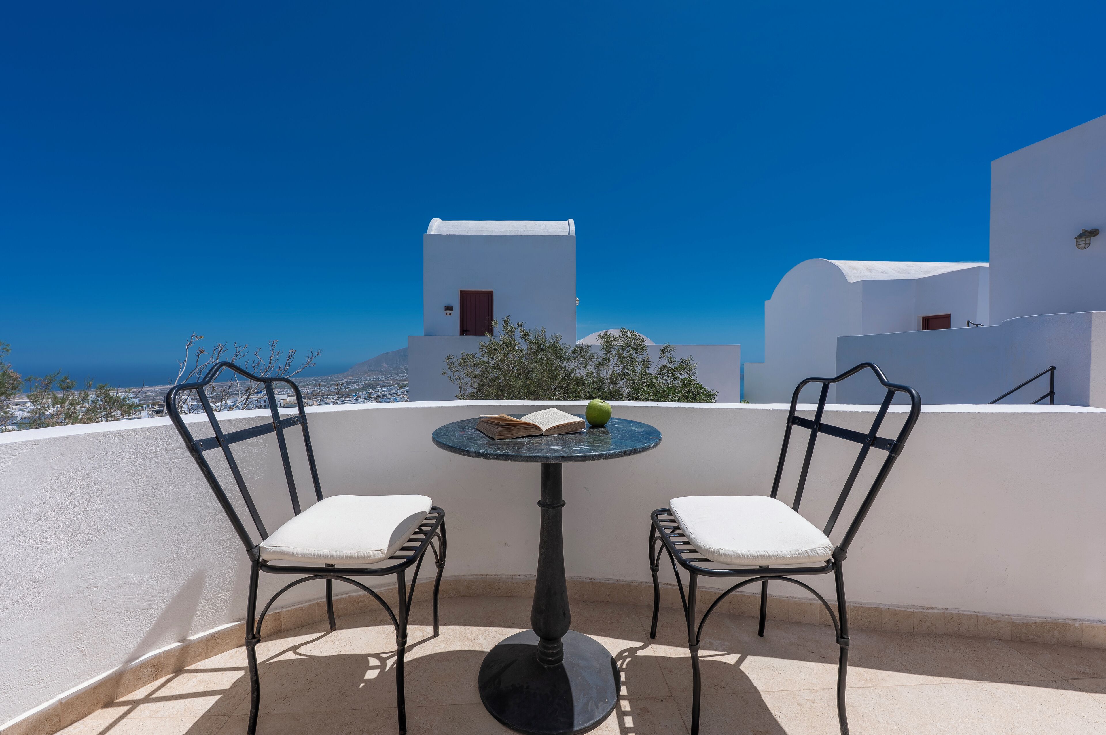 Une terrasse avec deux chaises et une table, offrant une vue sur la côte, avec des bâtiments blancs et un ciel bleu dégagé.
