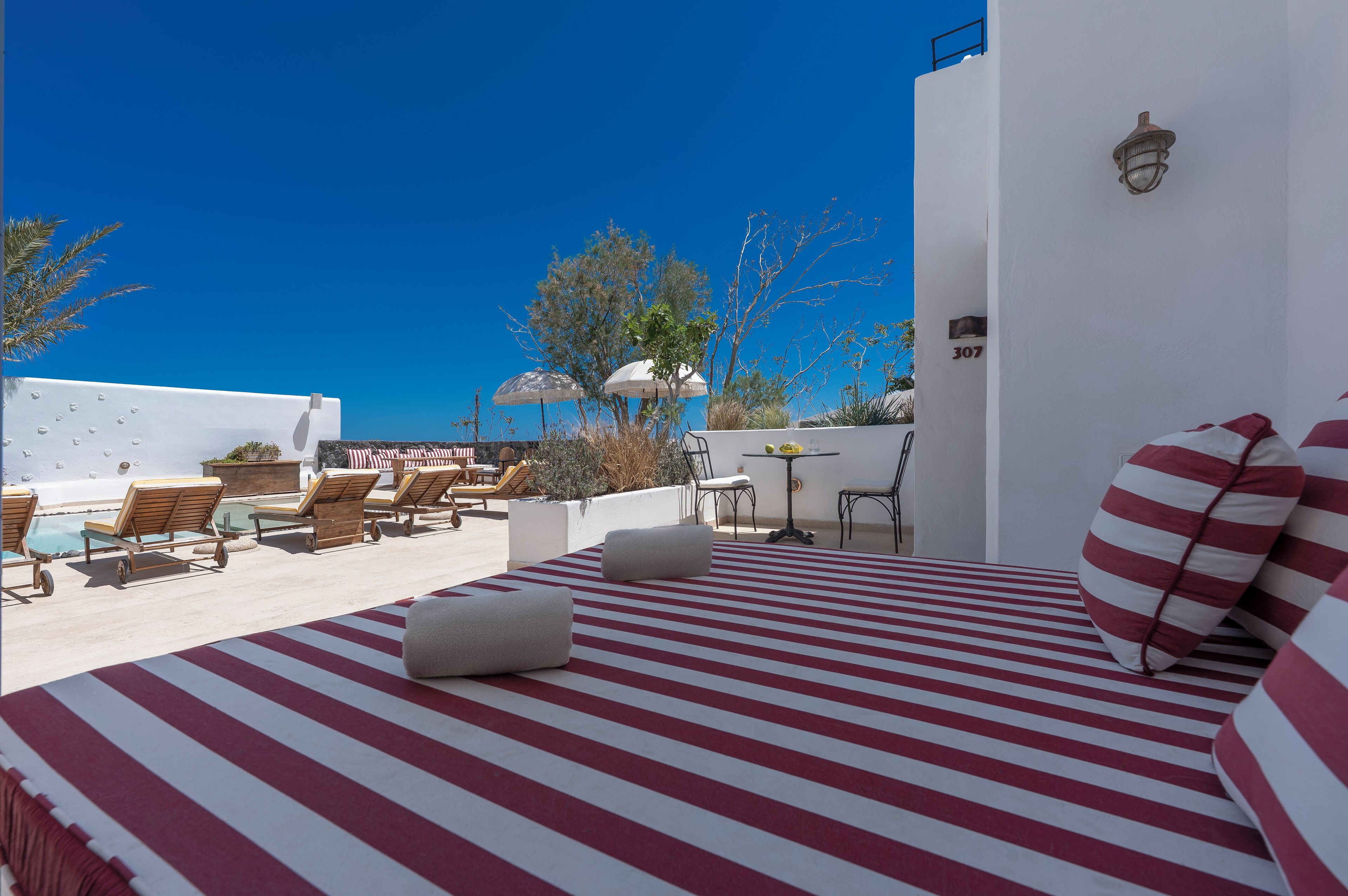 A hotel room with a large bed, a pool, lounge chairs, and a view of the sea.