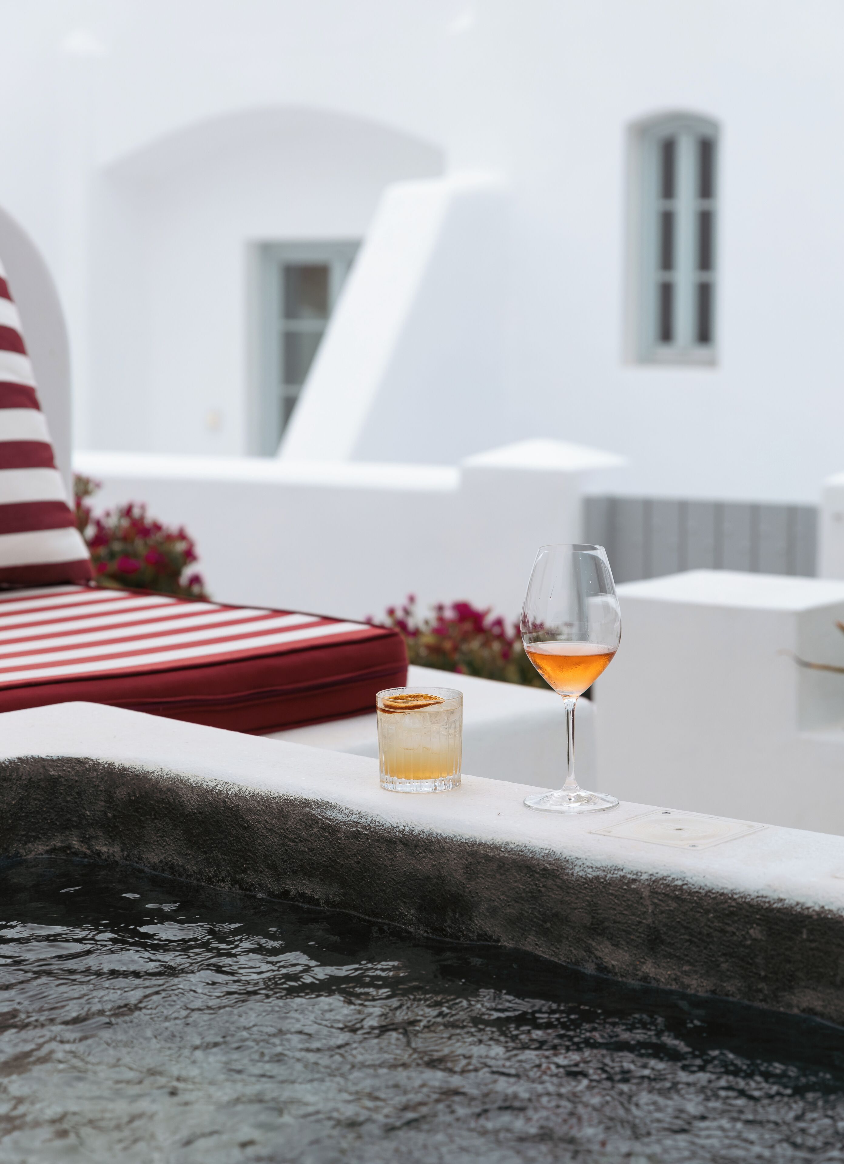 A glass of amber-colored drink and a small glass with a slice of orange on a white surface, with a red-striped cushion and a red bag in the background.