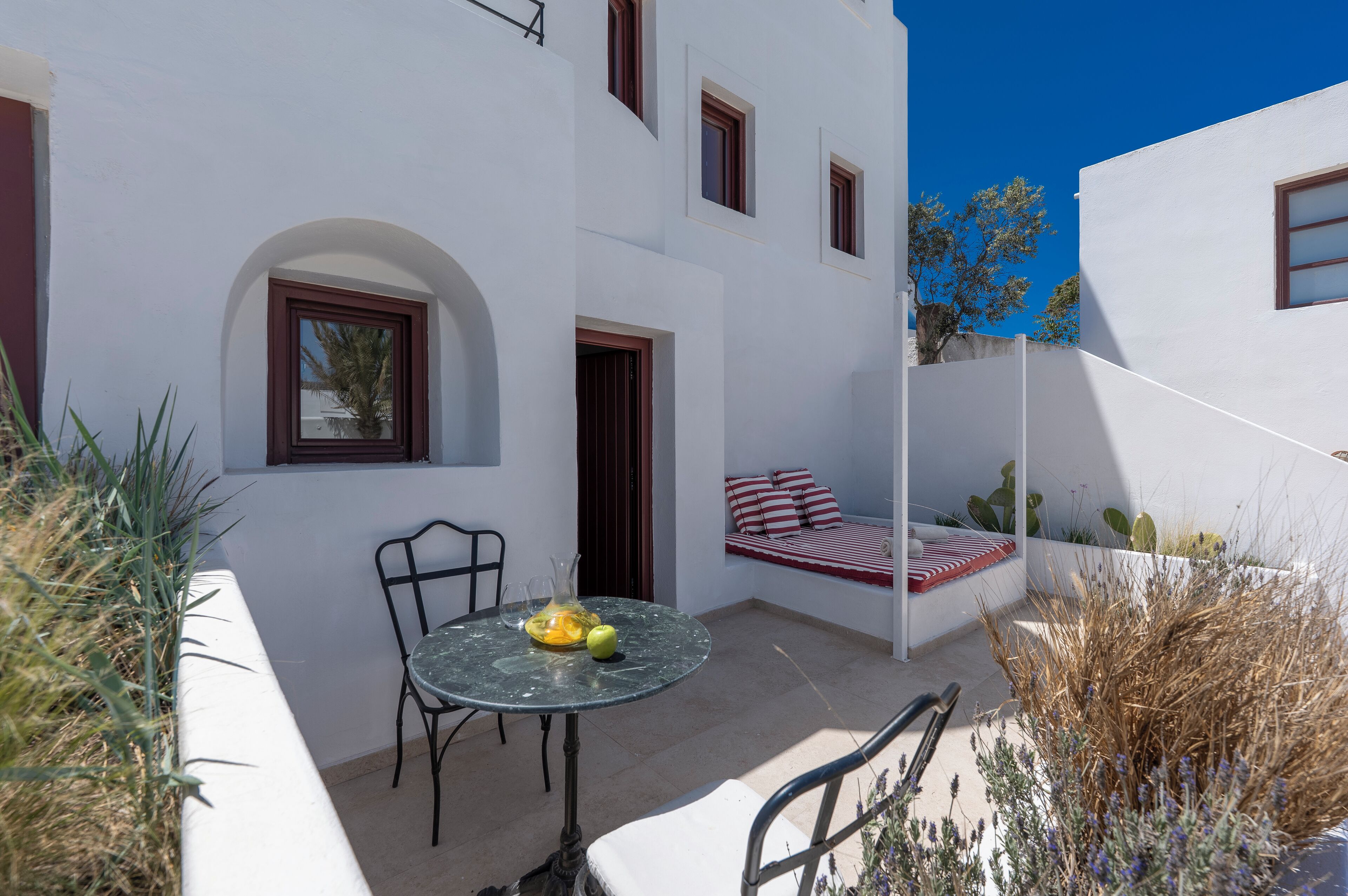 Un edificio blanco con un pequeño patio que cuenta con una mesa redonda, dos sillas y un banco acolchado con cojines a rayas rojas y blancas.