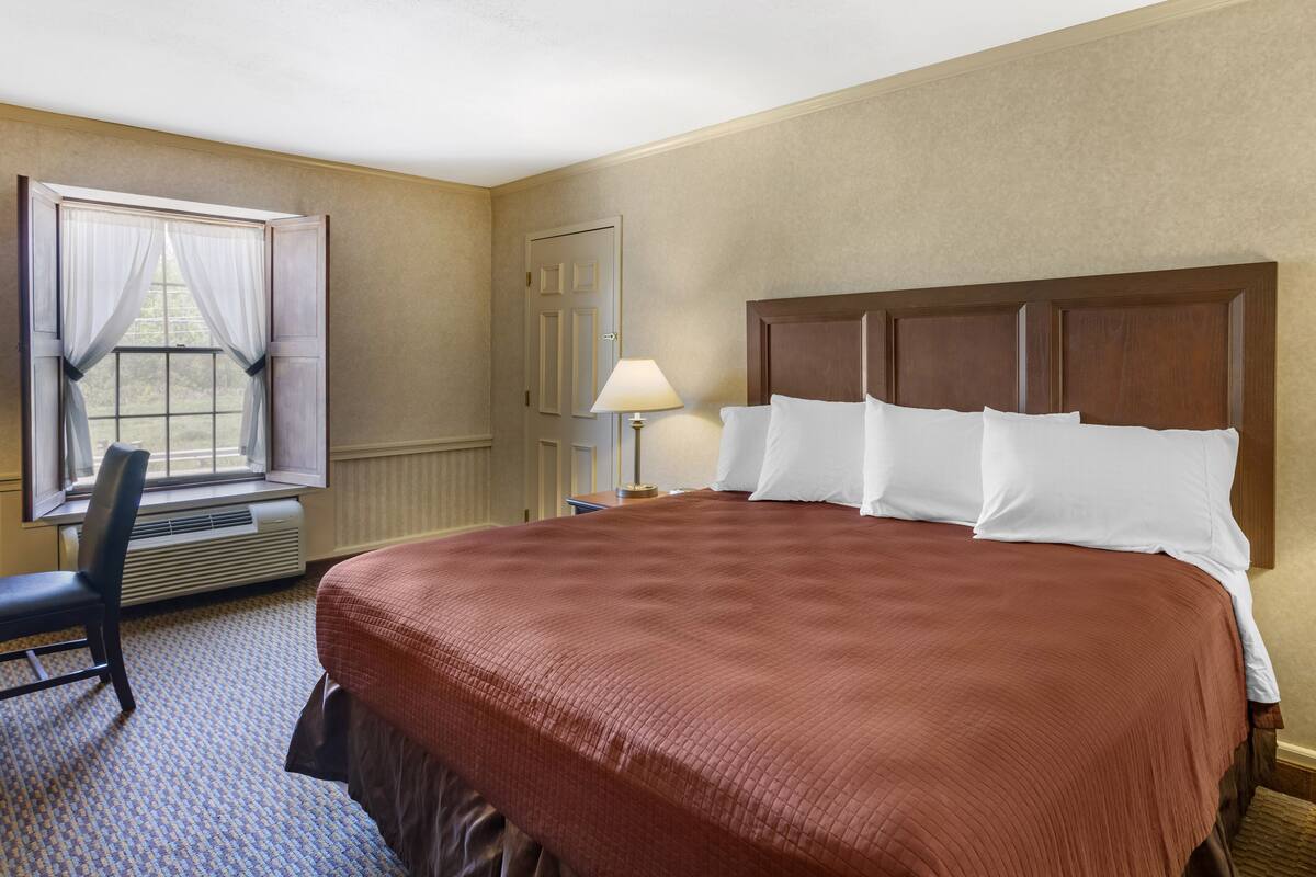 Quarto de hotel com uma cama grande, uma cadeira, uma janela com cortinas, uma luminária e uma porta.