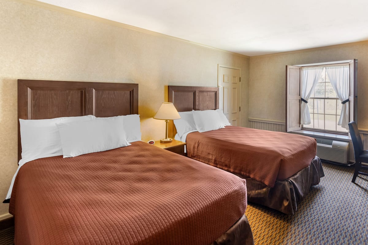 Quarto de hotel com duas camas, uma mesa de cabeceira com abajur, uma janela com cortinas e uma cadeira.