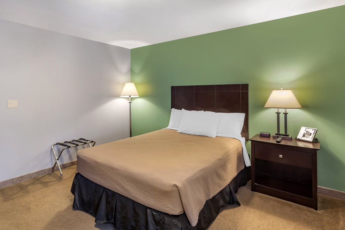 Cama bem arrumada com colcha marrom e saia preta, duas luminárias de cabeceira, mesa de cabeceira com telefone e porta-retrato, e um pequeno suporte de metal na parede.