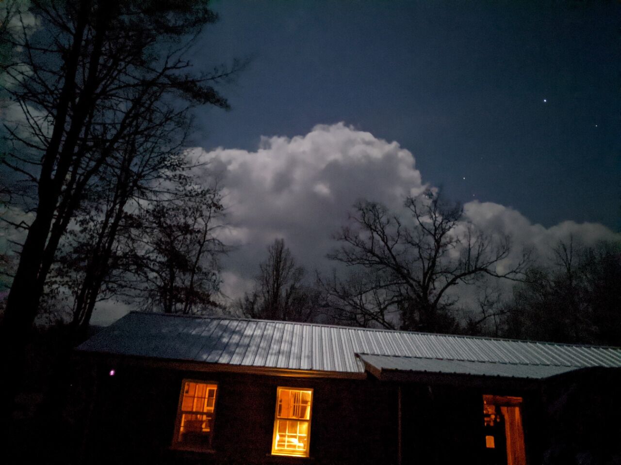 A house with a metal roof and two lit windows, surrounded by trees and a cloudy sky.