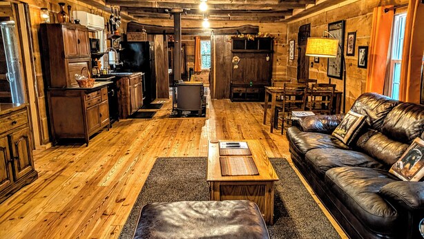 A rustic cabin interior with a wooden floor, a leather sofa, a dining table, and a kitchen area with wooden cabinets and a refrigerator. - Cozy 1 - Bedroom Cabin in Cooper's Creek Area (Suches)