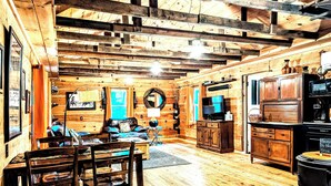 A rustic cabin interior with a wooden dining table, chairs, a sofa, a television, and a kitchen area with a stove. - Cozy 1 - Bedroom Cabin in Cooper's Creek Area (Suches)
