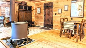 A rustic wooden cabin interior with a central wood-burning stove, a wooden bench, a dining table with chairs, and framed pictures on the walls. - Cozy 1 - Bedroom Cabin in Cooper's Creek Area (Suches)