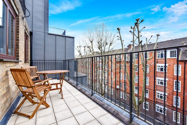 A balcony with a table and chairs, overlooking a brick building and trees.