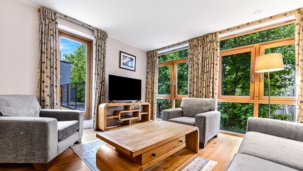 Ein modernes Wohnzimmer mit einem Flachbildfernseher, zwei grauen Sofas, einem Holztisch und großen Fenstern mit Vorhängen.