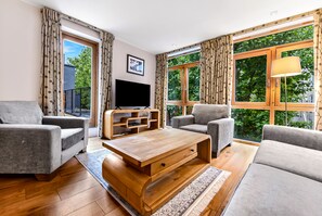 A modern living room with a flat-screen TV, two grey sofas, a wooden coffee table, and large windows with curtains.