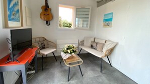 Un espace de vie compact comprenant une télévision, une guitare, un fauteuil en osier, un canapé moelleux et une petite table agrémentée d’un arrangement floral.