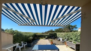 Un espace extérieur aménagé en terrasse, avec un auvent à rayures bleues et blanches, une table entourée de chaises et un banc.
