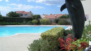 Une piscine ornée d’une statue de dauphin, entourée de verdure et avec des maisons en arrière-plan.