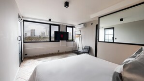 Una habitación de hotel moderna con una cama grande, un televisor montado en la pared y vistas a la ciudad a través de amplios ventanales.