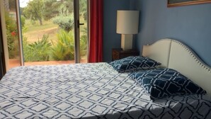 A bedroom with a patterned bedspread, a nightstand with a lamp, and a view of greenery through a sliding door. - Pieds dans l'eau Secret Creek (Conca)