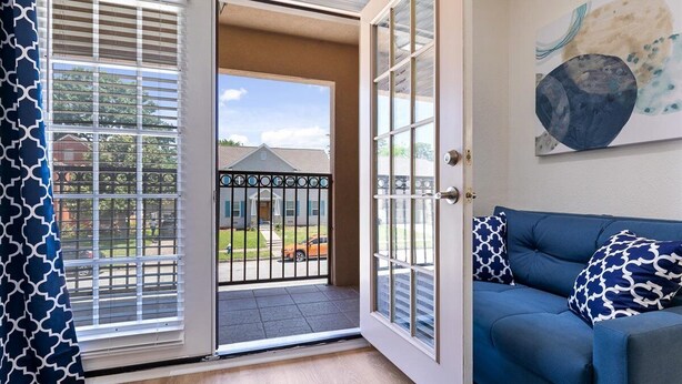 A room with a blue sofa, a large abstract painting, and a sliding glass door leading to a balcony with a view of a house and a fence. - Get Together & Stay-Play-Relax! Peaceful Oasis w/ TVs, Games, & BBQ -LUCKY House (Houston)