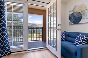 Une pièce avec un canapé bleu, un grand tableau abstrait et une porte-fenêtre coulissante donnant sur un balcon d’où l’on peut admirer une maison et une clôture.