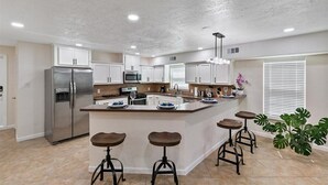 A modern kitchen with a central island, stainless steel refrigerator, white cabinets, and bar stools. - Get Together & Stay-Play-Relax! Peaceful Oasis w/ TVs, Games, & BBQ -LUCKY House (Houston)