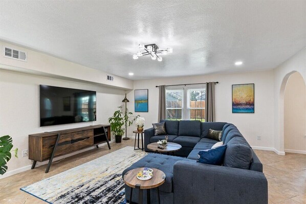 A modern living room with a sectional sofa, a flat-screen TV, a wooden console, and a large window. - Get Together & Stay-Play-Relax! Peaceful Oasis w/ TVs, Games, & BBQ -LUCKY House (Houston)