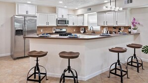 A modern kitchen with white cabinets, a stainless steel refrigerator, a microwave, and a large island with stools. - Get Together & Stay-Play-Relax! Peaceful Oasis w/ TVs, Games, & BBQ -LUCKY House (Houston)
