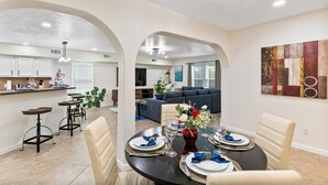A dining area with a round table set for four, a kitchen in the background, and a living room with a sofa and TV. - Get Together & Stay-Play-Relax! Peaceful Oasis w/ TVs, Games, & BBQ -LUCKY House (Houston)
