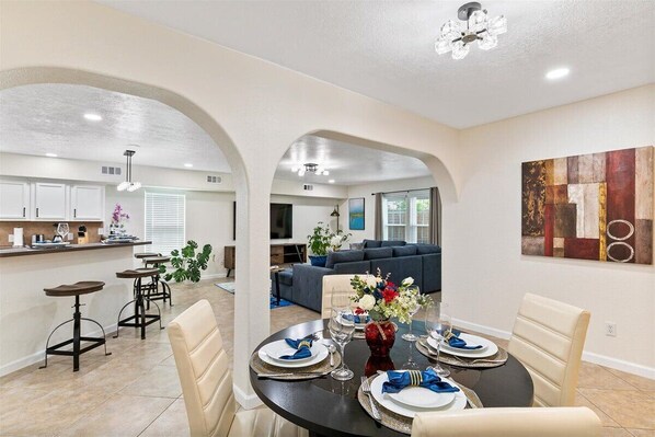 A dining area with a round table set for four, a kitchen in the background, and a living room with a sofa and TV.