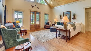 A spacious living room with a high ceiling, wooden flooring, and a large window. There are two doors, a sofa, a chair, and a table with a lamp. - Spacious Midway Cottage w/ Porches, Near Hunting! (Midway)