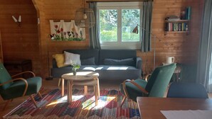 A cozy living room with a sofa, two chairs, a small table, a patterned rug, a window with curtains, a hanging light, and a bookshelf.