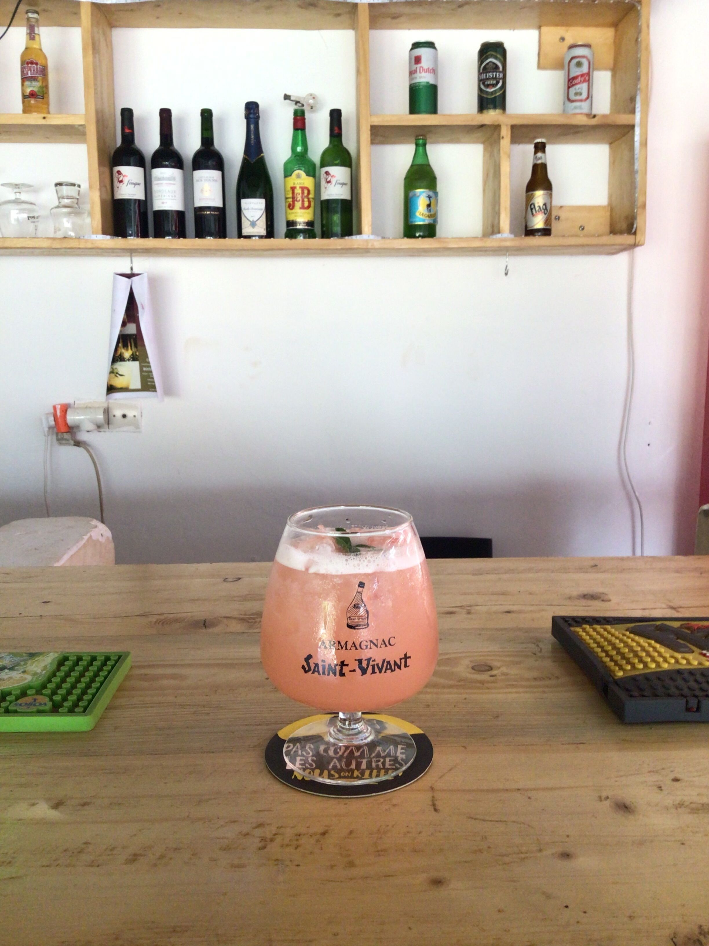 Lasillinen Saint-Vivant-cocktailia puupöydällä, jonka päällä on näppäimistö ja alusta. Taustalla on hylly, jossa on erilaisia pulloja, ja pieni kyltti.