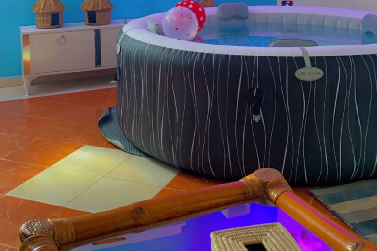 A room with a large round hot tub, a flat-screen TV mounted on the wall, a small white cabinet, and a wall-mounted shelf with decorative items.