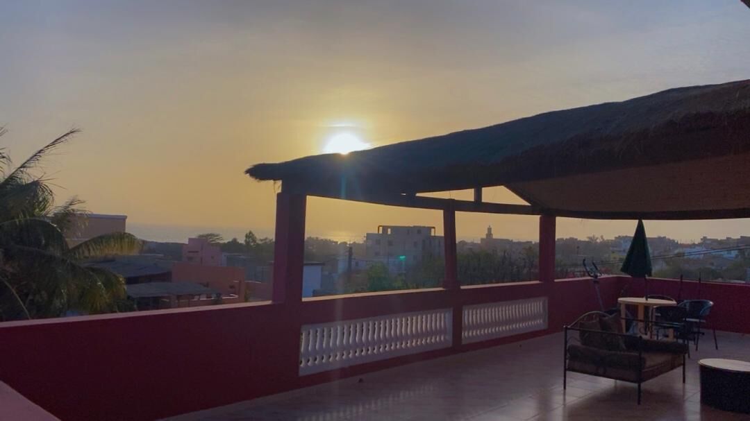 A balcony with a view of the sunset, a thatched roof, and a seating area with chairs and a table.