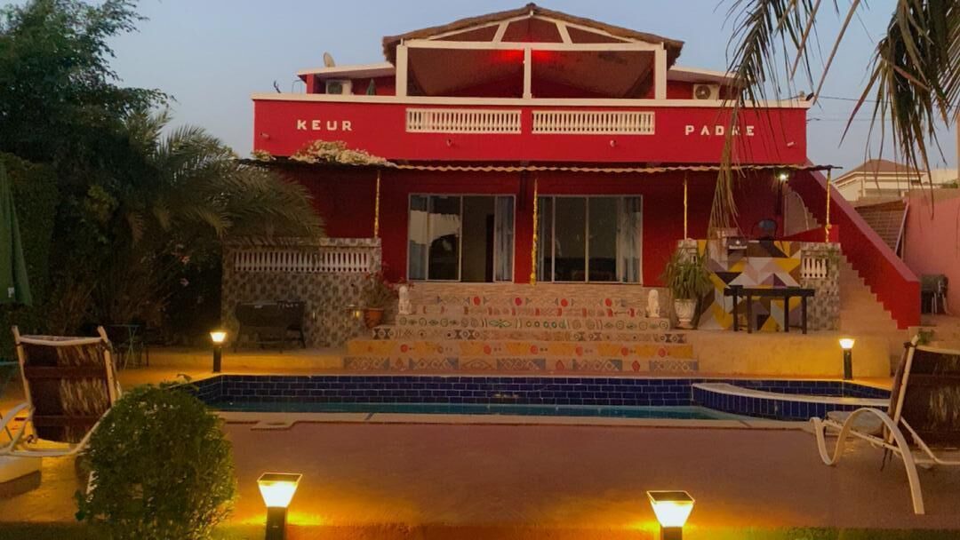 A red building with 'KEUR PADRE' on the facade, a pool, and outdoor seating.