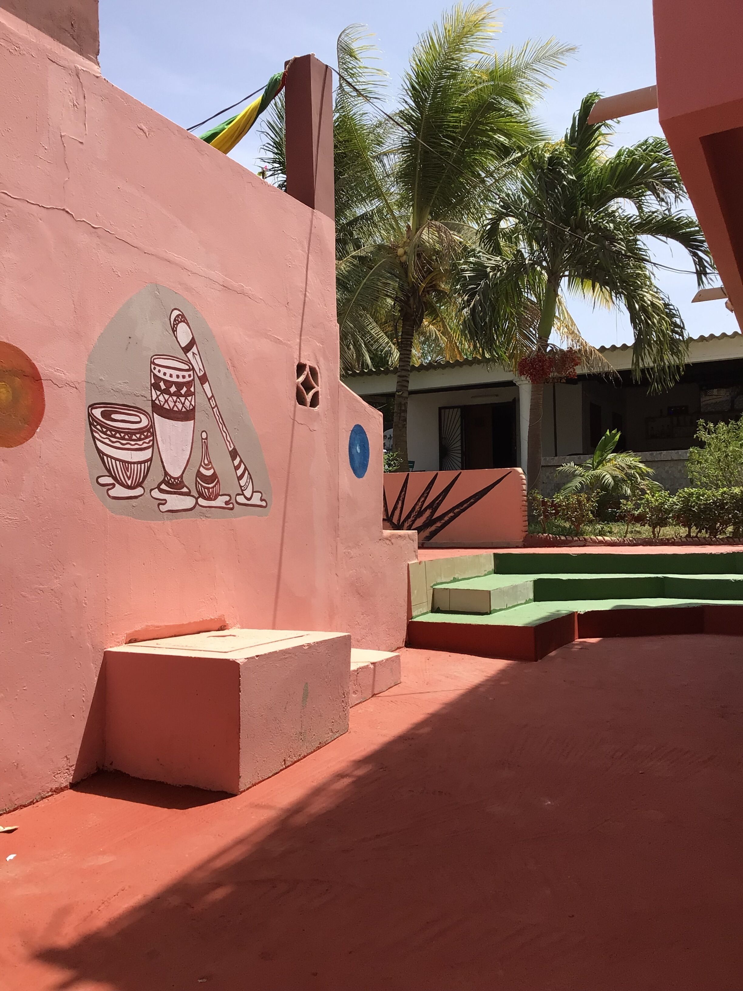 A building with a pink wall featuring a mural of traditional pottery and a blue circular design. There are steps leading up to the building, and palm trees are visible in the background.