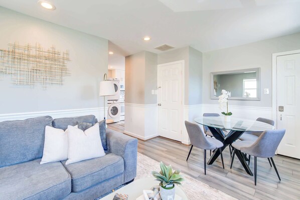 A modern living room with a grey sofa, a glass dining table with chairs, and a kitchen area with a washing machine.
