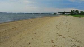 A sandy beach with a calm sea, a few boats in the distance, and houses along the shoreline.
